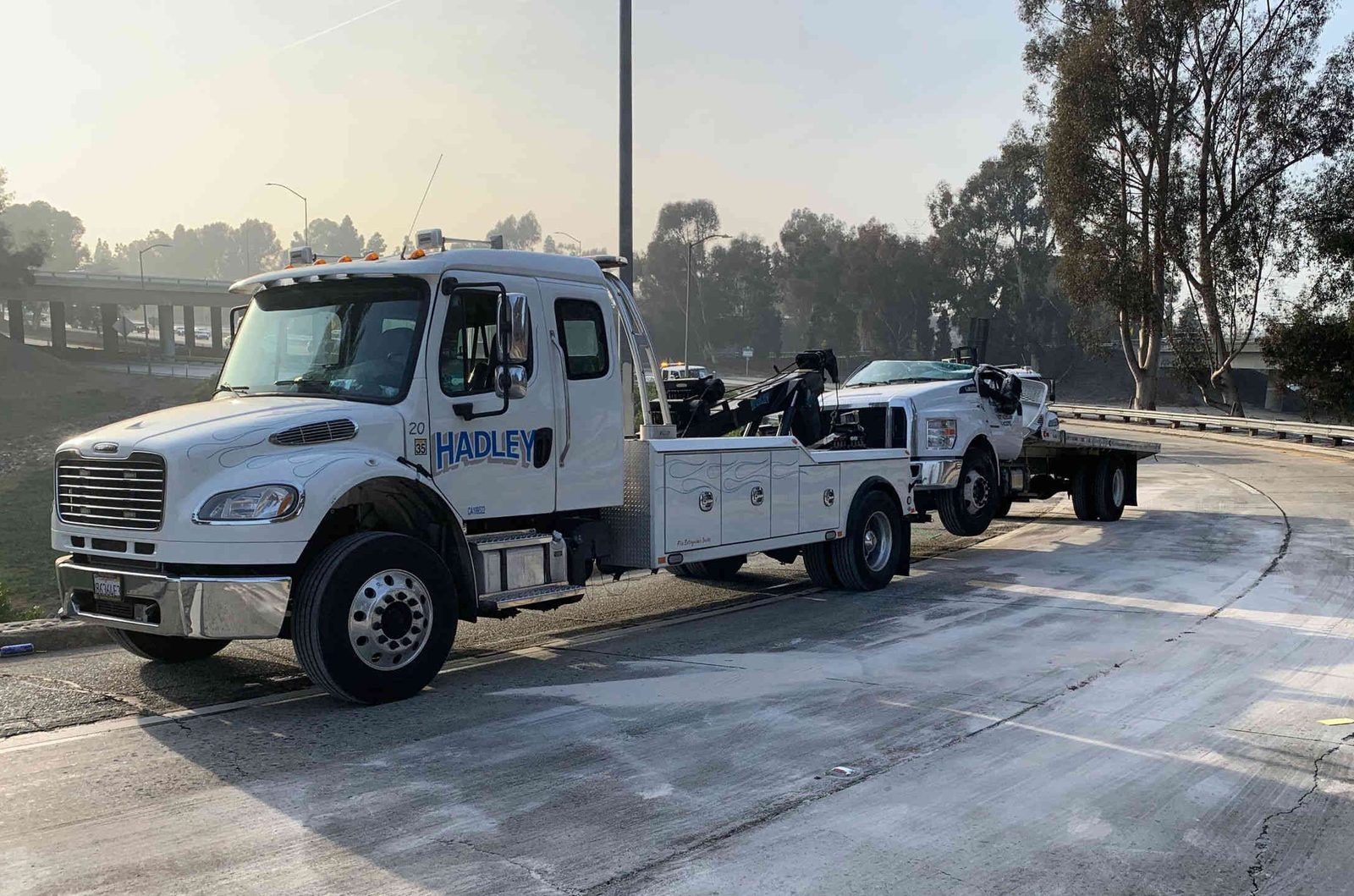 Hadley medium-duty tow truck pulling a disabled flatbed on a SoCal freeway ramp